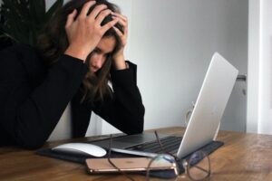 A dark-haired woman sites in front of a laptop computer, hands on her head. She's clearly frustrated.