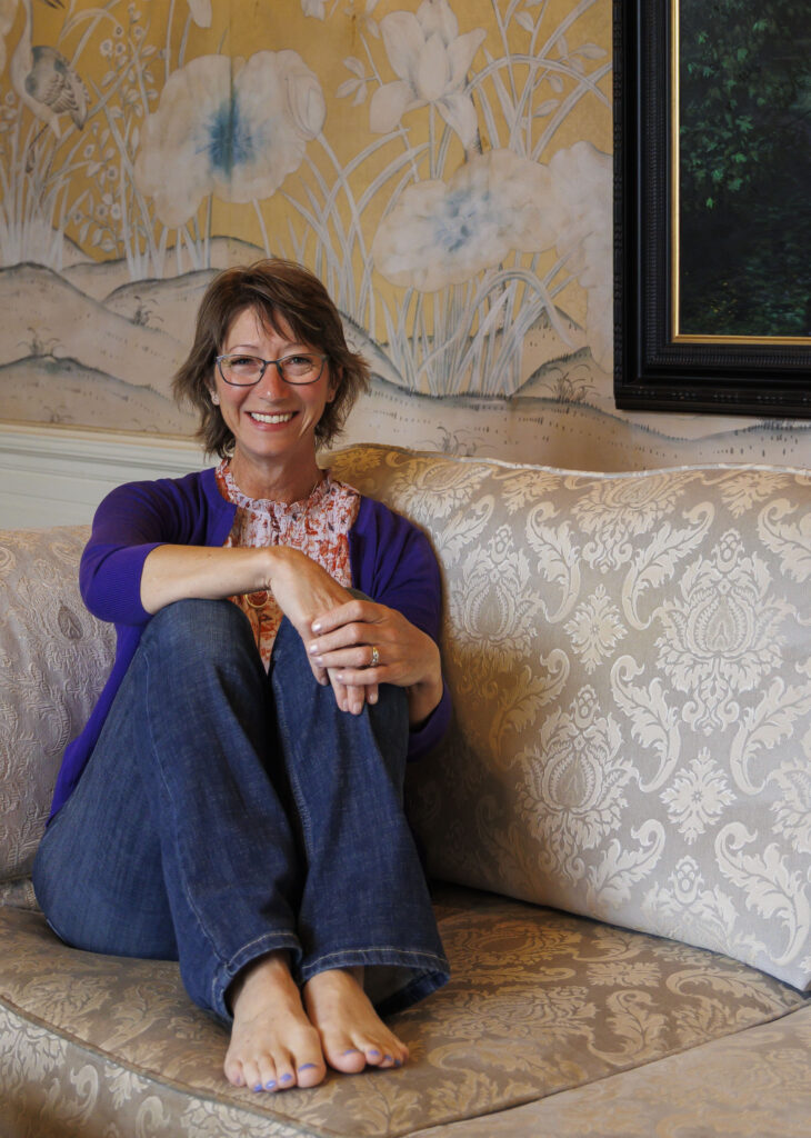 A women short hair sits on a couch with bare feet and her hands are on her bended knees.